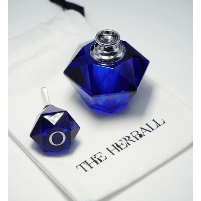 A blue perfume bottle leaning on a white pouch with The Herball logo on a white surface.