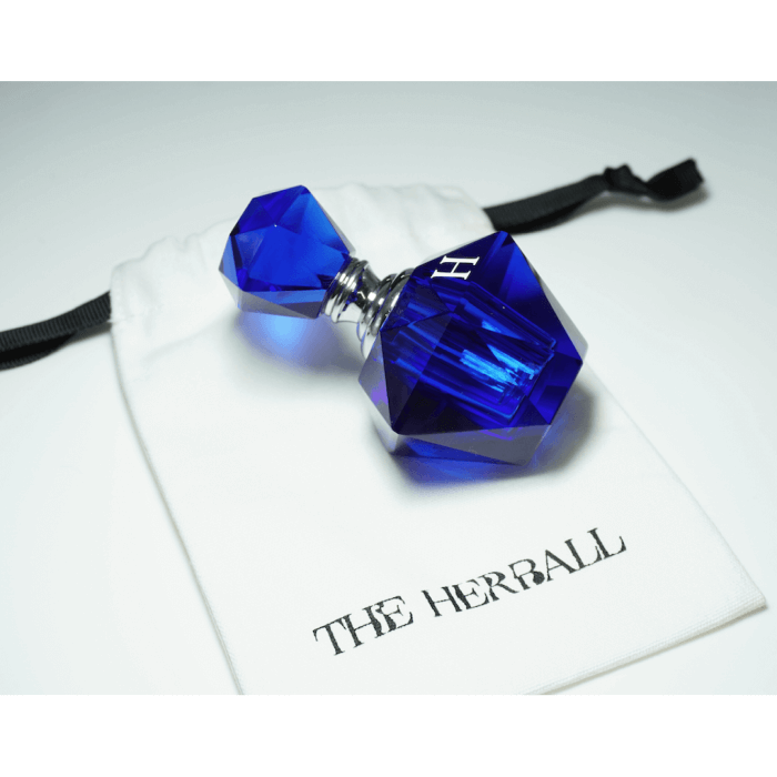 A blue perfume bottle leaning on a white pouch with The Herball logo on a white surface.